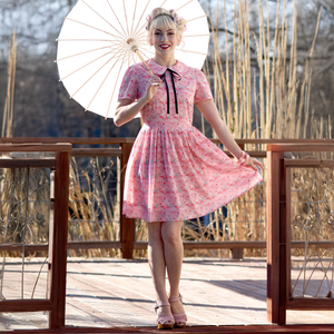 A woman wearing a pink dress with a flamingo print, featuring a Peter Pan collar and short sleeves, standing with a parasol. The dress has a mid-century design.
